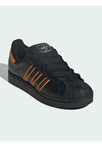 Baskets noires Adidas avec un nez en caoutchouc texturé, rayures orange, onglet de talon assorti et lacets noirs sur un fond clair.