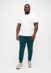 Witte katoenen t-shirt met rode logo, gecombineerd met teal joggers en grijze sneakers. De outfit heeft een casual pasvorm en een gladde textuur.
