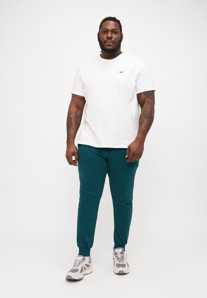 Witte katoenen t-shirt met rode logo, gecombineerd met teal joggers en grijze sneakers. De outfit heeft een casual pasvorm en een gladde textuur.
