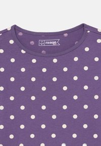 Μωβ βαμβακερό μπλουζάκι με στρογγυλή λαιμόκοψη και λευκές πουά που είναι τοποθετημένες ομοιόμορφα σε όλο το ύφασμα. Η ετικέτα είναι ορατή μέσα στον γιακά.