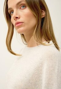 Jasnobeżowy sweter z dzianiny o okrągłym dekolcie, miękkiej fakturze i delikatnym prążkowaniu wzdłuż szwów, noszony przez modelkę z długimi włosami.