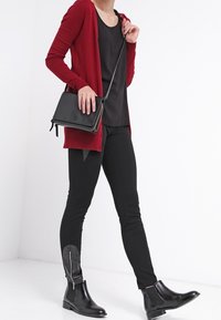 Rote Strickjacke, schwarzes Oberteil, schwarz enganliegende Hose mit Reißverschlussdetails. Schwarze Knöchelstiefel. Schlichte Umhängetasche aus dunkelbraunem Leder. Einheitlicher weißer Hintergrund.