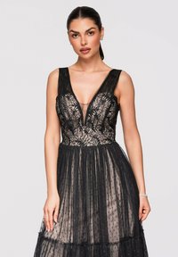 Femme aux cheveux foncés en queue de cheval lisse porte une robe noire en dentelle sans manches avec une jupe transparente à pois, debout sur un fond clair.
