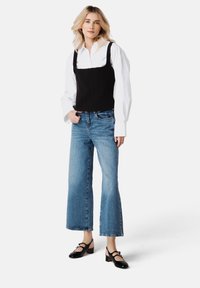 Top sans manches noir, chemise blanche à boutons, jean large bleu clair, et chaussures à talons noires avec des brides autour de la cheville. Design simple et moderne.