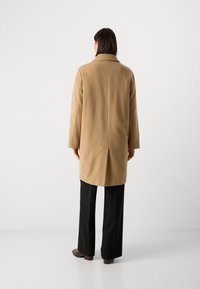 Manteau tan au design structuré, avec un grand col, des manches longues et une fente à l'arrière ; associé à un pantalon noir.