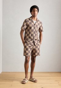 Rövid ujjú gombos ing és hozzáillő sort, geometrikus barna és bézs mintázattal. Fekete szandálban viselve. Simább anyagtextúra.