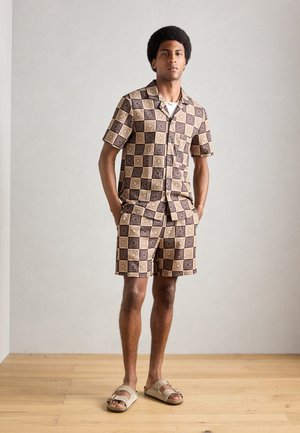 Trumpas rankovės mygtukų marškiniai ir derančios šortai geometriniu rudu ir smėlio raštu. Dėvimi su smėliniais šlepetėmis. Lygus audinio paviršius.