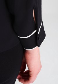 Poignet de chemise noire à manches longues avec un passepoil blanc, fermeture à bouton, et une fente laissant voir un peu de poignet sur une main à la peau claire, suspendue de manière détendue.