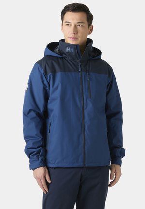 Uomo che indossa una giacca impermeabile blu navy con cappuccio, in piedi con le mani rilassate, che guarda leggermente di lato.