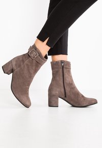 Bottines en suède taupe avec un talon épais, équipées d'une fermeture éclair sur le côté et d'une sangle décorative à boucle autour de la cheville.