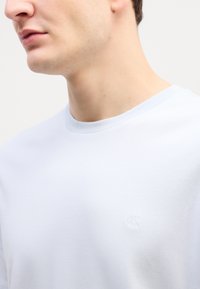 Bílý bavlněný tričko s kulatým výstřihem a krátkými rukávy, s jemně vyšitým logem na hrudi, hladká textura.
