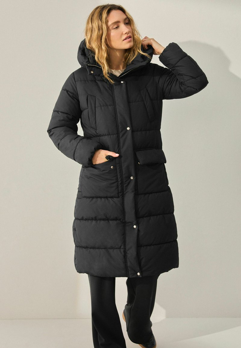 Femme dans un long manteau rembourré noir avec capuche, ajustant ses cheveux, se tenant devant un fond clair.