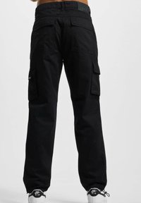 Pantalons cargo noirs en coton, dotés de poches latérales et arrière, de poignets élastiques et d'une texture lisse. Associés à des baskets blanches.