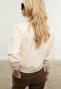 Cremefarbener Sweater mit gerippten Bündchen und Saum, kombiniert mit braunen Hosen. Die Haare sind lang und wellig, und die Figur wird von hinten gezeigt.