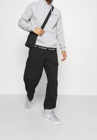 Grijze hoodie, zwarte cargobroek met merklogo op de tailleband en grijze sneakers. Materiaal lijkt een textuur te hebben, casual pasvorm met zijzakken.