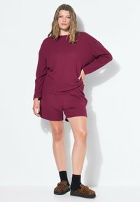 Mujer con una camiseta de manga larga suelta de color burdeos con pantalones cortos a juego y zapatos de leopardo slip-on con calcetines negros.
