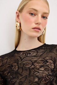 Top in pizzo nero con intricati motivi floreali e paisley, dotato di scollatura rotonda e una texture trasparente. Grandi orecchini ad anello dorati accentuano il look.