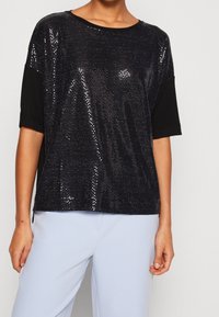 Černý top s krátkým rukávem, který má na přední straně flitry a na zadní straně jednobarevnou černou látku, pohodlný střih, kulatý výstřih a boční rozparky.