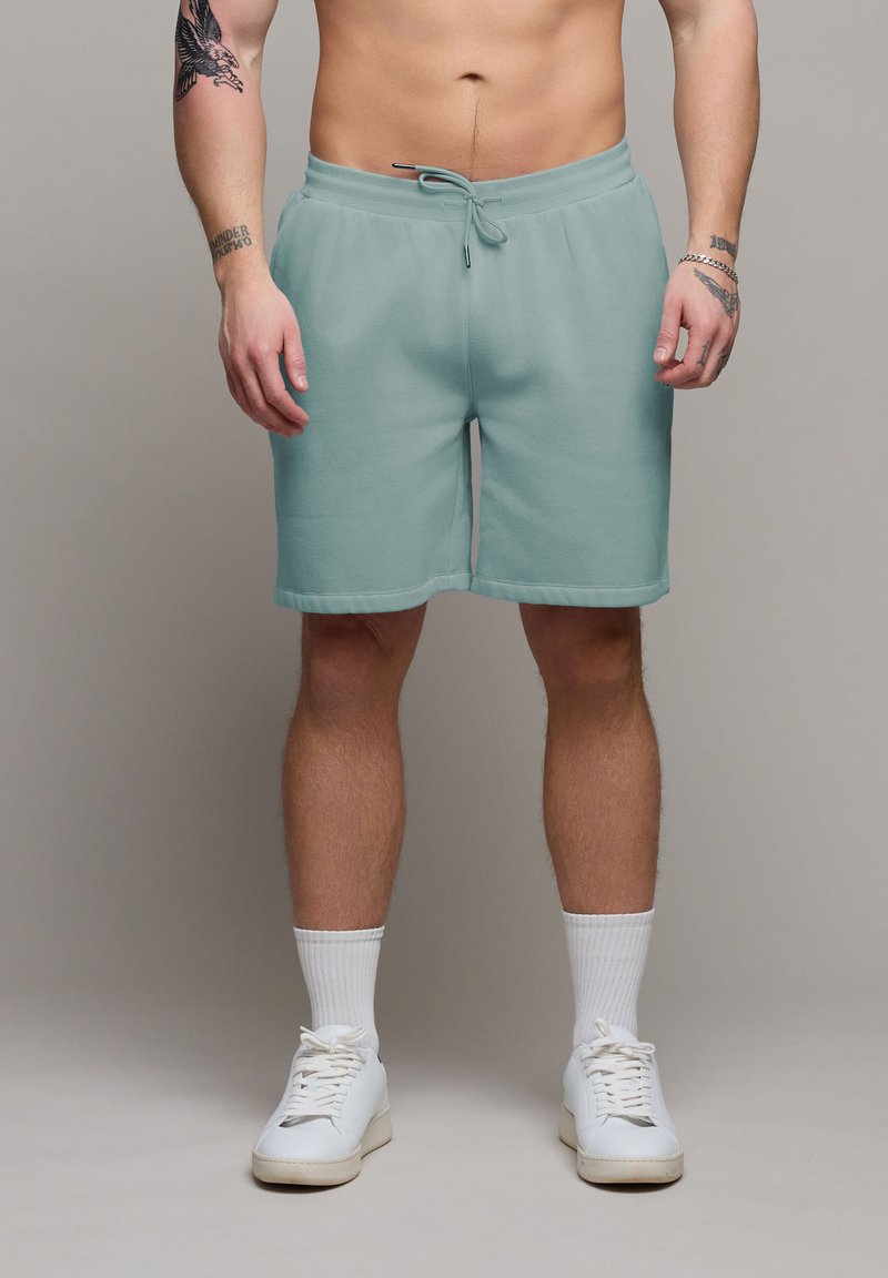 Homme portant un short vert clair à cordon de serrage, des chaussettes blanches hautes et des baskets blanches, montrant des tatouages sur les bras sur un fond neutre.