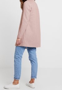 Cardigan lungo rosa chiaro con taglio dritto, tessuto strutturato, tasche laterali, abbinato a jeans azzurri e sneakers bianche.