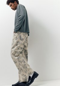 Homme en pantalon beige à motifs, chemise grise à manches longues et chaussures noires, debout de profil avec les mains dans les poches, sur fond blanc.