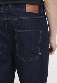 Jeans de mezclilla azul oscuro con un parche de cuero marrón en la cinturilla y costuras en contraste. Incluye dos bolsillos traseros con una etiqueta.
