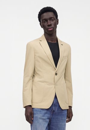 Joven con una americana beige sobre una camisa negra y vaqueros azules, de pie frente a un fondo blanco liso.