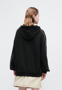 Osoba s valovitom kosom do ramena nosi crni oversized duksericu s bijelim šavovima na rukavima i bež suknju, okrenuta leđima.