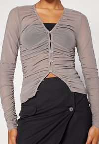 Hellgraue durchsichtige Bluse mit langen Ärmeln, gerafftem Material, V-Ausschnitt und vorderen Knöpfen, kombiniert mit einem schwarzen Rock mit einer Seitentasche.