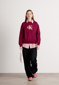 Bordový sveter s logom 'Calvin Klein', vrstvený nad svetloružovou pruhovanou košeľou, čiernymi širokými nohavicami a ružovými topánkami.