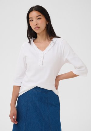 Femme aux longs cheveux foncés portant un chemisier blanc à col à volants et une jupe en jean bleue, main sur la hanche, debout devant un fond clair.