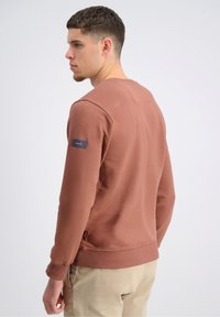 Felpa marrone con polsini e orlo a costine, caratterizzata da una piccola etichetta con logo sulla manica sinistra e un design a collo rotondo. Realizzata in tessuto testurizzato.