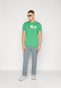 Grønn t-skjorte med kort erme og grafisk design på forsiden, sammen med lysegrå bukser og grå joggesko. Tilbehør inkluderer hvite solbriller.