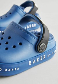 Plave klompe od teksturiranog sintetičkog materijala, s crnim podesivim remenom i bijelim natpisom "BAKER" sa strane.
