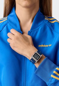 Kék adidas cipzáros dzseki sárga csíkokkal, egy kézre húzva, amely egy ezüst színű digitális órát mutat, kék szíjas és digitális kijelzővel.
