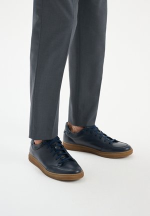 Navy leather sneakers with a textured finish, round toe, and rubber sole. Features navy laces and minimal branding. Paired with gray trousers.