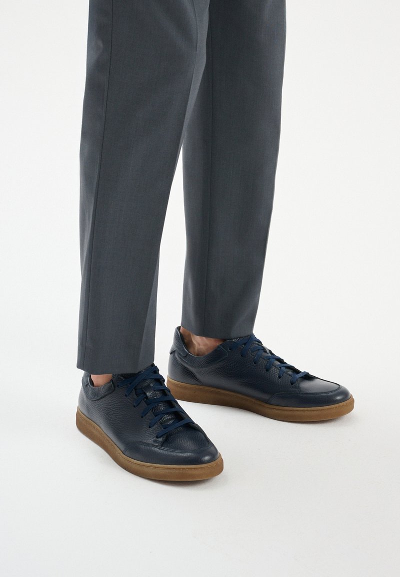 Tenisi din piele navy cu un finisaj texturat, vârf rotund și talpă din cauciuc. Prezintă șireturi navy și un branding minimalist. Asortați cu pantaloni gri.