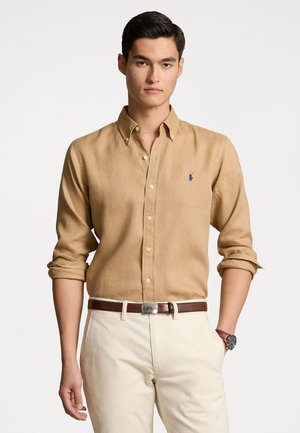 Camicia button-up marrone chiaro con maniche arrotolate, dotata di un piccolo logo blu sul petto, abbinata a pantaloni color crema e una cintura marrone.