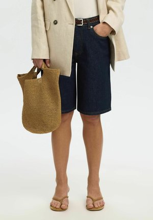 Persona che indossa un blazer beige, un top bianco, pantaloncini di jeans scuri fino al ginocchio, sandali color cuoio, tenendo una borsa tote marrone intrecciata di lato.