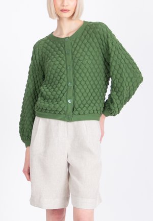 Kvinde iført en textureret grøn cardigan med knapper og puffede ærmer samt højttaljede lyse beige shorts mod en hvid baggrund.