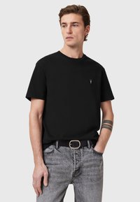 T-shirt in cotone nero con scollatura rotonda, maniche corte, con un piccolo logo ricamato sul petto, abbinato a jeans grigi chiari.