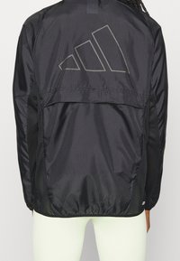 Czarna kurtka windbreaker o teksturowanym wykończeniu, z dużym szarym logo z tyłu, siateczkowymi panelami bocznymi i elastycznymi mankietami.