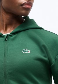 Κοντινό πλάνο ενός ατόμου που φοράει πράσινο φούτερ με φερμουάρ, με ένα μικρό κεντητό λογότυπο κροκοδείλου στο στήθος.