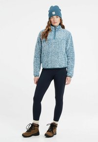 Pullover blu chiaro in pile con zip a quarti, abbinato a leggings blu navy e stivaletti da trekking marroni. Completato da un berretto di maglia blu.