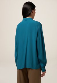 Bluză teal cu guler, mâneci lungi și croială lejeră. Textură netedă și luciu subtil, asortată cu pantaloni maro.