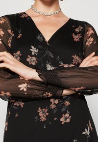 Vestido negro con estampado floral, de mangas largas y transparentes adornadas con flores rosas y blancas; presenta un escote en forma de V y puños recogidos.