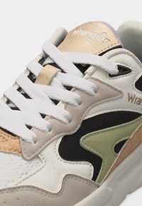 Αθλητικό παπούτσι με λευκά κορδόνια, μπεζ και ελιές χρωματισμούς, υφή λεπτομερειών και το λογότυπο Wrangler στη γλώσσα και στο πλαϊνό τμήμα.