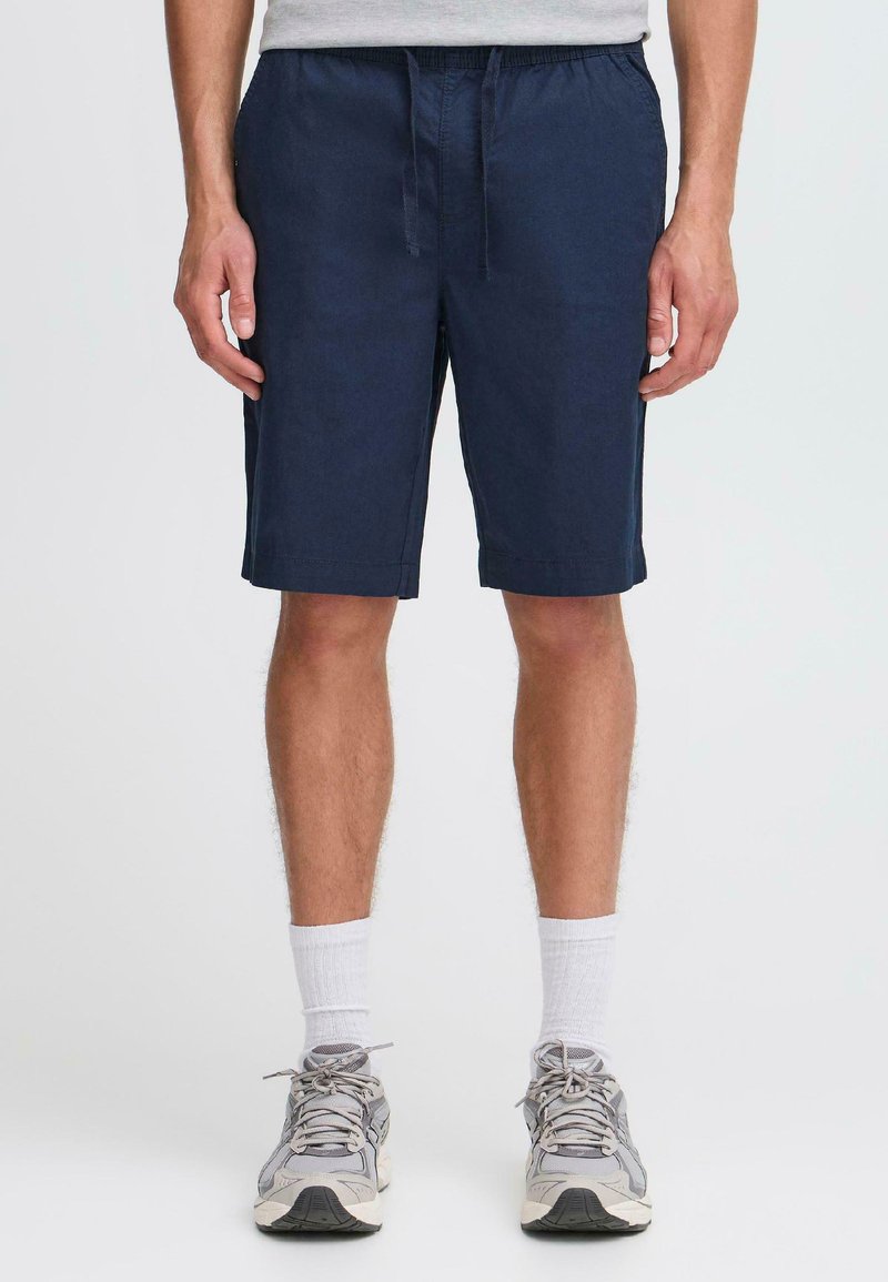 Shorts bleu marine en tissu léger avec une ceinture élastique et des poches sur les côtés ; associés à des chaussures de sport grises et des chaussettes blanches.