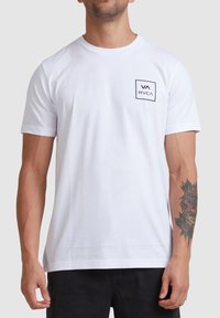 T-shirt en coton blanc avec des manches courtes et un col rond. Présente un petit logo carré noir sur la poitrine gauche et une texture lisse.
