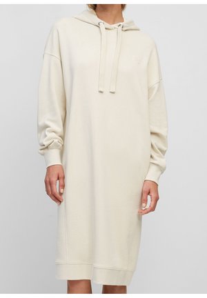 Frau trägt ein lockeres, cremefarbenes Kapuzensweatshirtkleid mit langen Ärmeln und Kordelzugkapuze, stehend vor einem schlichten Hintergrund.